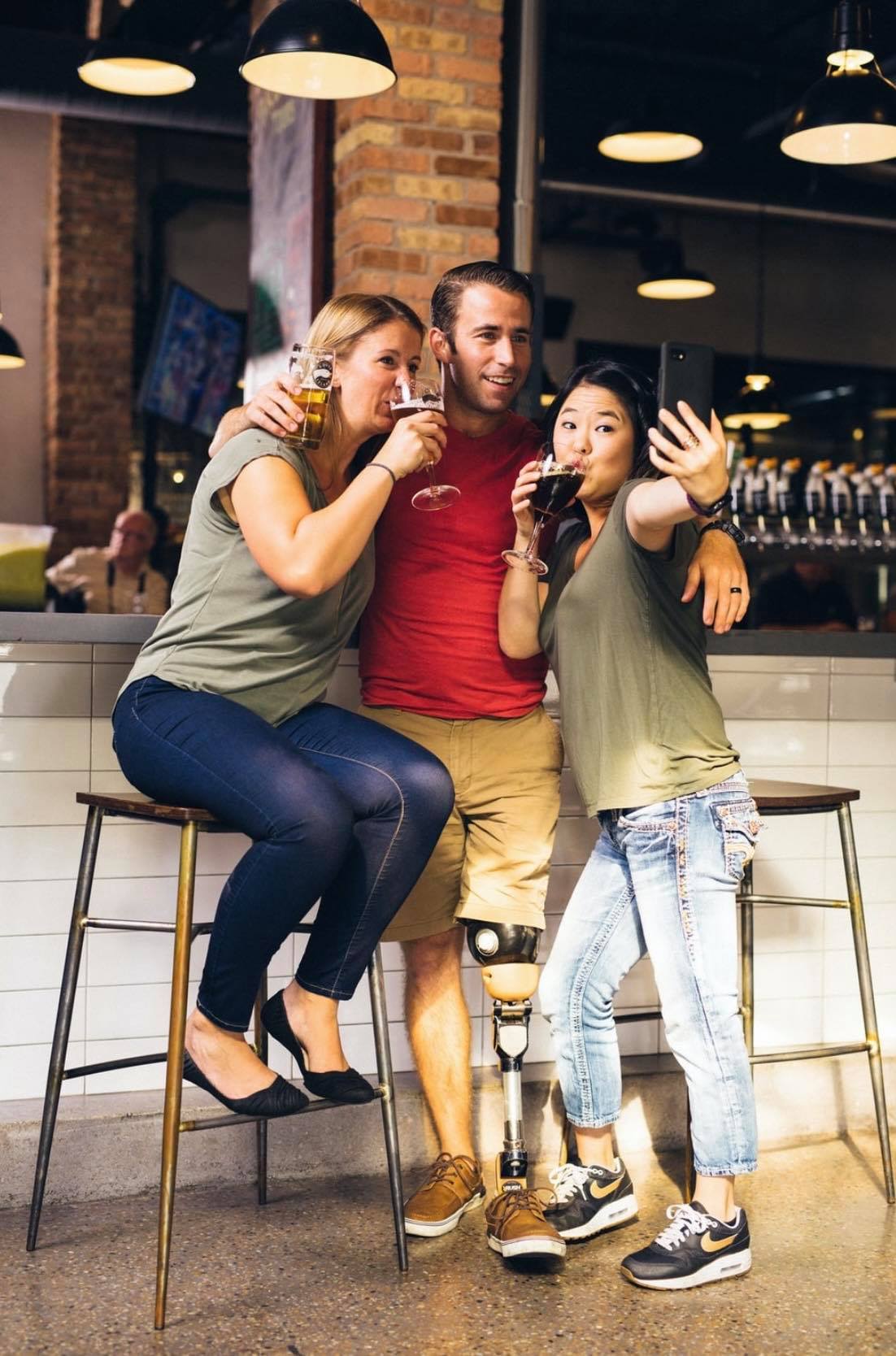 Image appears to be of three people in a bar taking a group selfie. There is a man standing in the middle holding his drink, arms around the shoulders loosely of the two woman and he has a prosthetic leg. The woman on the left leans in posing with her drink up and the woman on the right leans into him pretending to drink and taking the photo with a phone.